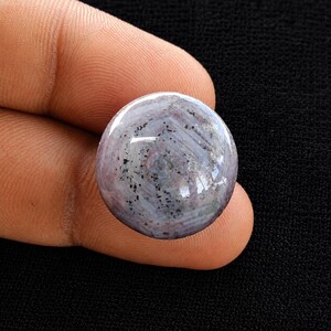 May include: A round, cabochon-cut gemstone featuring a translucent, pale lavender and grey base with a distinct six-rayed star pattern and scattered black mineral inclusions throughout the surface, displaying a smooth, polished, and reflective finish.