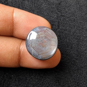 May include: A round, cabochon-cut gemstone featuring a translucent, greyish-blue body with a distinct, centred six-rayed star pattern. The stone displays subtle internal banding and a smooth, polished surface that reflects light across its dome.