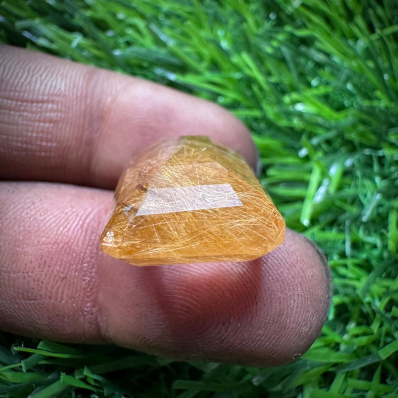 May include: A golden-orange gemstone, pyramid-shaped. The stone has a polished, reflective surface with visible internal structures. The gemstone is held in a person's hand, with a green grassy background.