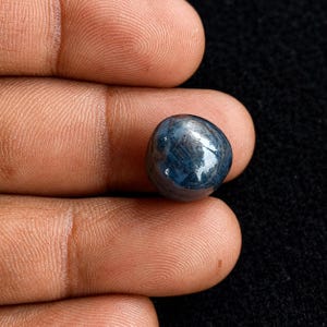 Op de afbeelding: Een ronde, in cabochon geslepen saffier edelsteen met een diepblauwe kleur en zichtbare interne insluitingen. De steen heeft een glad, gepolijst oppervlak dat licht reflecteert en een mix van donkere en lichtere blauwtinten laat zien.
