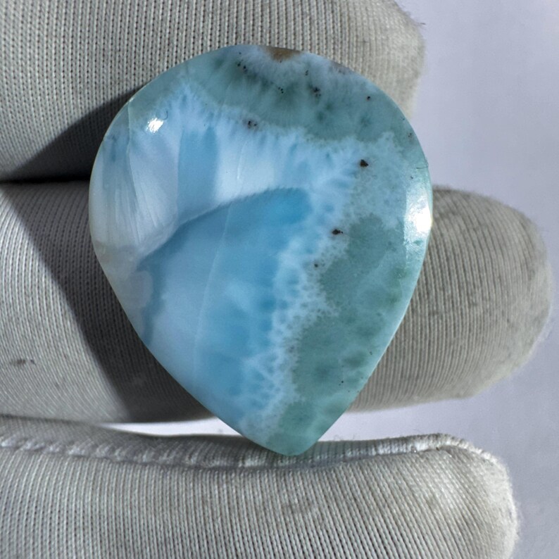 May include: A polished, teardrop-shaped gemstone with a swirling pattern of light and dark blue hues, resembling ocean waves. The stone has a smooth, glossy surface and is likely a piece of larimar, a rare gemstone.