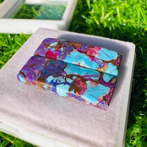 Puede incluir: Dos cabujones de piedras preciosas rectangulares con una vibrante mezcla de colores. Las piedras presentan una base turquesa con vetas moradas, rosas y doradas. Los cabujones se exhiben sobre una superficie blanca, con hierba verde al fondo.