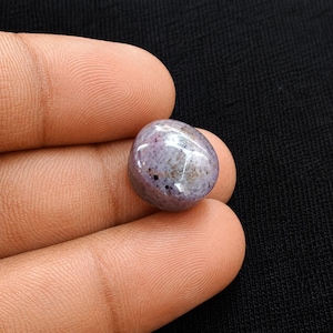 Peut inclure: Une pierre précieuse cabochon polie et lisse avec une forme arrondie en dôme. La pierre présente un aspect marbré avec des nuances de violet clair, de lavande et de gris, rehaussées par de petites taches noires réparties sur sa surface.