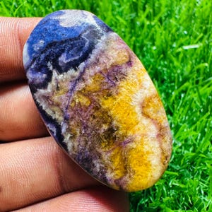 Op de afbeelding: Een ovale edelsteen met een gepolijst oppervlak. De steen heeft een mix van kleuren, waaronder diepblauw, paars, geel en wit. De kleuren zijn wervelend en hebben patronen, waardoor een unieke en natuurlijke uitstraling ontstaat. De edelsteen wordt in een hand gehouden.