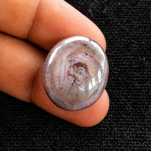 Puede incluir: Una piedra preciosa cabujón pulida de forma ovalada, con un tono marrón violáceo apagado, un patrón interno hexagonal distintivo y una superficie lisa y reflectante. La piedra muestra inclusiones naturales y sutiles variaciones de color en toda su extensión.