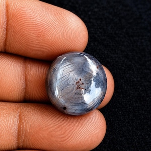 Op de afbeelding: Een ronde, in cabochon geslepen edelsteen met een donkerblauw-grijze kleur en een zichtbaar stervormig optisch effect. Het oppervlak vertoont een vezelige, chatoyante textuur met subtiele lichtreflecties en kleine, donkere roodbruine insluitingen.