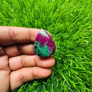 Könnte beinhalten: Ein tropfenförmiger, polierter Edelstein. Der Stein weist eine lebendige Farbmischung auf, darunter tiefes Rot, Smaragdgrün und Schwarz. Der Edelstein wird vor einem Hintergrund aus grünem Kunstgras gehalten.