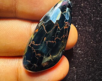 May include: A polished, elongated gemstone with a dark blue base and intricate orange and yellow veining. The stone's surface reflects light, showcasing a mix of colors and patterns. The gemstone is held between fingers.