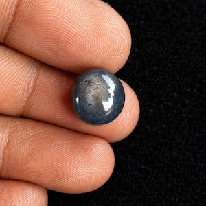 Può includere: Una piccola pietra preziosa rotonda tagliata a cabochon, di colore blu-grigio scuro con una superficie liscia e lucida. La pietra presenta un sottile riflesso sulla parte superiore bombata, mostrando una tonalità profonda e tenue.