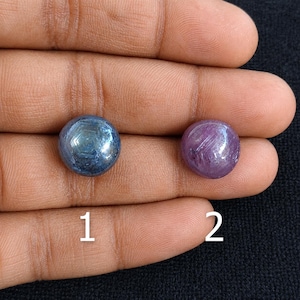 Peut inclure: Deux pierres précieuses cabochons polies et rondes. La pierre 1 est d'un bleu profond avec un motif en étoile hexagonal visible. La pierre 2 est d'un violet vif avec des inclusions cristallines internes. Les deux sont numérotées 1 et 2.
