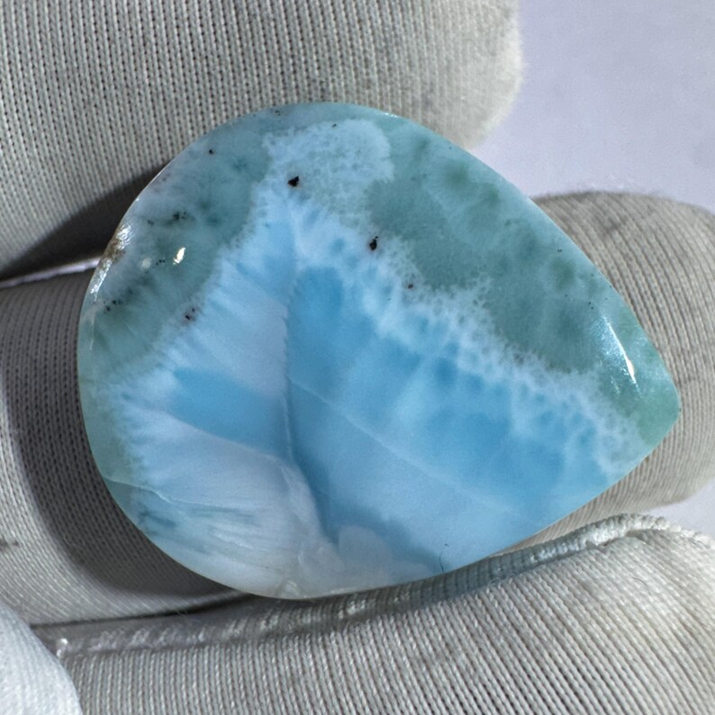 May include: A polished, teardrop-shaped larimar gemstone. The stone features a blend of light blue, white, and seafoam green hues, with a marbled appearance. Small dark inclusions are scattered across the surface, adding detail to the natural stone.