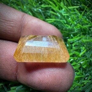 May include: A faceted, golden-yellow gemstone with internal golden inclusions. The gemstone is held between fingers against a backdrop of green grass. The cut of the stone is rectangular with angled sides.