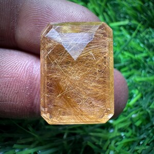 May include: A rectangular, faceted gemstone with a golden-brown hue. The stone has internal golden needle-like inclusions, creating a unique visual texture. The gem is held up against a green background.