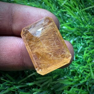 May include: A rectangular, faceted gemstone with a golden-yellow hue and internal needle-like inclusions. The gem is held against a backdrop of green grass, showcasing its clarity and colour. The stone's cut reveals multiple facets.