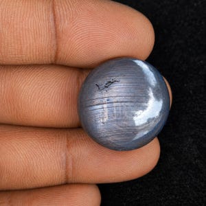 Peut inclure: Une pierre précieuse ronde taillée en cabochon avec une surface lisse et polie, d'une couleur bleu-gris métallique foncé. La pierre présente des stries horizontales visibles et une petite inclusion sombre en surface, reflétant la lumière.
