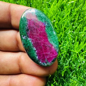 May include: An oval-shaped gemstone with a vibrant mix of colors. The stone features a striking magenta center, surrounded by a rich green hue. The polished surface reflects light, highlighting the natural patterns and textures of the stone.