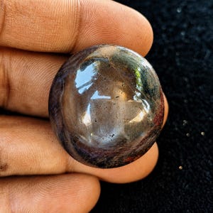 May include: A round, polished cabochon gemstone featuring a deep, dark brown and black color palette with subtle hints of blue and red. The surface is smooth and reflective, displaying natural inclusions and a glossy, high-shine finish.
