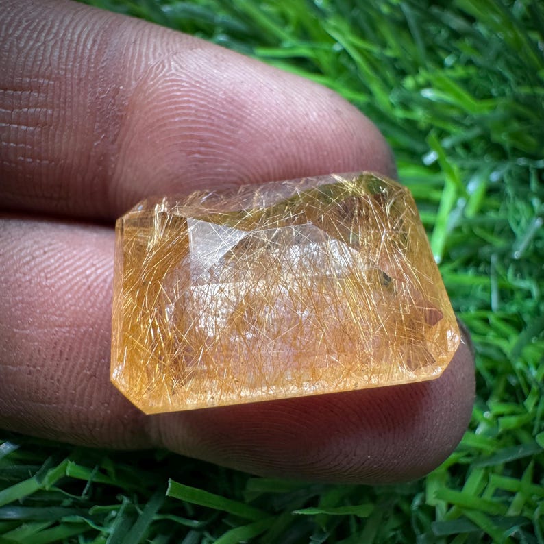 May include: A rectangular gemstone with a golden-yellow colour and internal needle-like inclusions. The gem is held between fingers against a backdrop of green grass. The stone's cut reveals multiple facets.