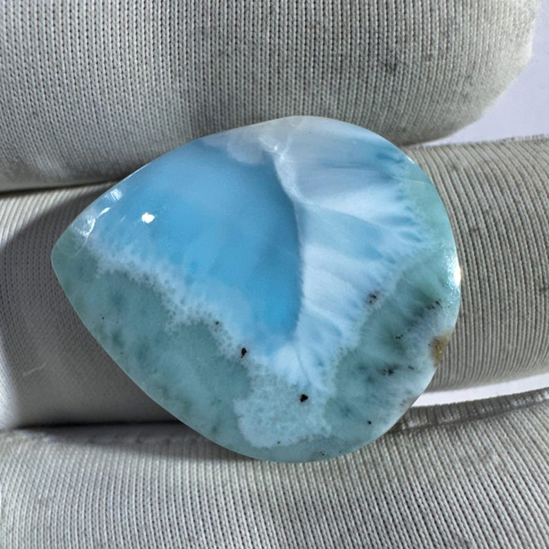 May include: A polished, teardrop-shaped gemstone with a light blue and white pattern. The stone has a smooth surface with areas of darker blue and small black inclusions. The overall appearance is reminiscent of ocean waves.