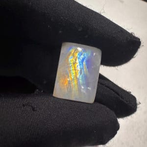 以下が含まれることがあります： 長方形の虹色のムーンストーン。石は青、黄色、オレンジの色合いで色の遊びを示しています。宝石は黒い手袋をはめた手で持たれており、石の鮮やかな色を際立たせています。