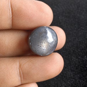 Peut inclure: Une pierre précieuse ronde taillée en cabochon avec une surface lisse et polie. La pierre présente une couleur gris foncé métallique avec des inclusions internes visibles et un éclat subtil et réfléchissant sur son sommet bombé.