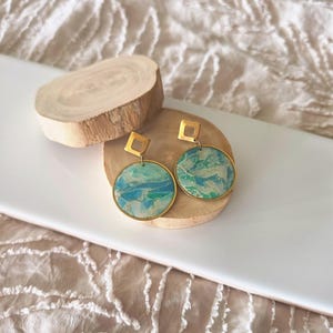 May include: A pair of drop earrings featuring gold-toned square posts connected to circular pendants. The circular pendants have a marbled pattern in shades of teal, blue, and white, encased in a thin gold-coloured metal rim.