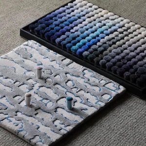 May include: A rectangular black tray containing rows of small, circular, tufted fabric samples in a gradient of blue, gray, and white tones. Beside it lies a square textile swatch featuring a textured, abstract gray and white pattern.