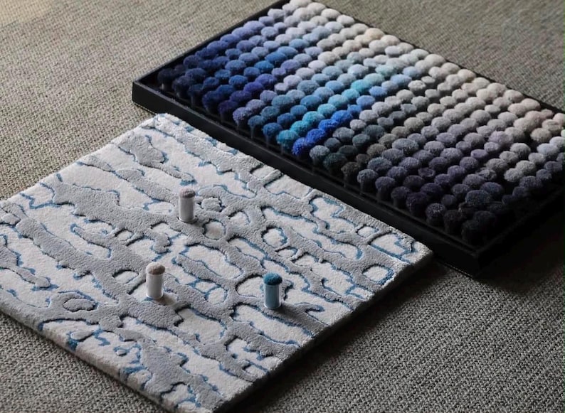 May include: A rectangular black tray containing rows of small, circular, tufted fabric samples in a gradient of blue, gray, and white tones. Beside it lies a square textile swatch featuring a textured, abstract gray and white pattern.