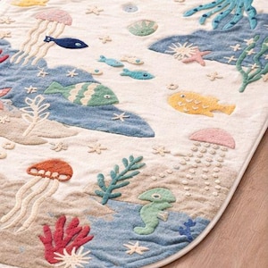 May include: An oval-shaped rug with a whimsical underwater scene. The rug features embroidered sea creatures like jellyfish, fish, and starfish in various colors, including blue, orange, and yellow. The background is a mix of beige and blue, creating a playful, decorative floor covering.