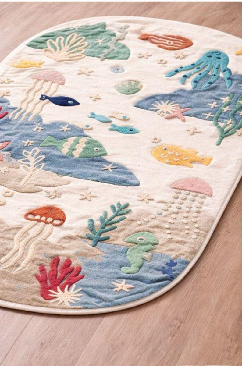 May include: An oval-shaped rug with a whimsical underwater scene. The rug features embroidered sea creatures like jellyfish, fish, and starfish in various colors, including blue, orange, and yellow. The background is a mix of beige and blue, creating a playful, decorative floor covering.