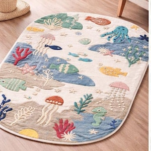 May include: An oval-shaped rug with a sea life design. The rug features colorful embroidered sea creatures, including fish, jellyfish, and an octopus, set against a cream-colored background with blue and beige accents. The rug is approximately 60 inches long.
