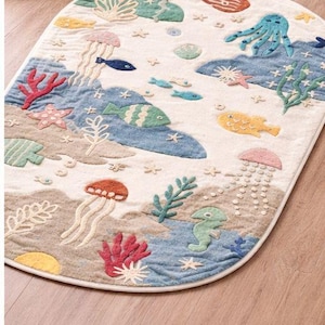 May include: An oval-shaped rug with an underwater scene. The rug features embroidered sea creatures like fish, jellyfish, and an octopus in shades of blue, green, red, and yellow. The background is a mix of beige and blue.