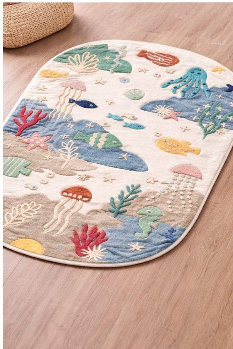 May include: An oval-shaped rug with an underwater scene. The rug features embroidered sea creatures like fish, jellyfish, and an octopus in shades of blue, green, red, and yellow. The background is a mix of beige and blue.