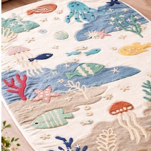 May include: An oval-shaped rug with a sea life design. The rug features various marine creatures, including fish, jellyfish, and an octopus, in shades of blue, green, orange, and yellow. The background is a sandy beige color, with blue water and white accents.