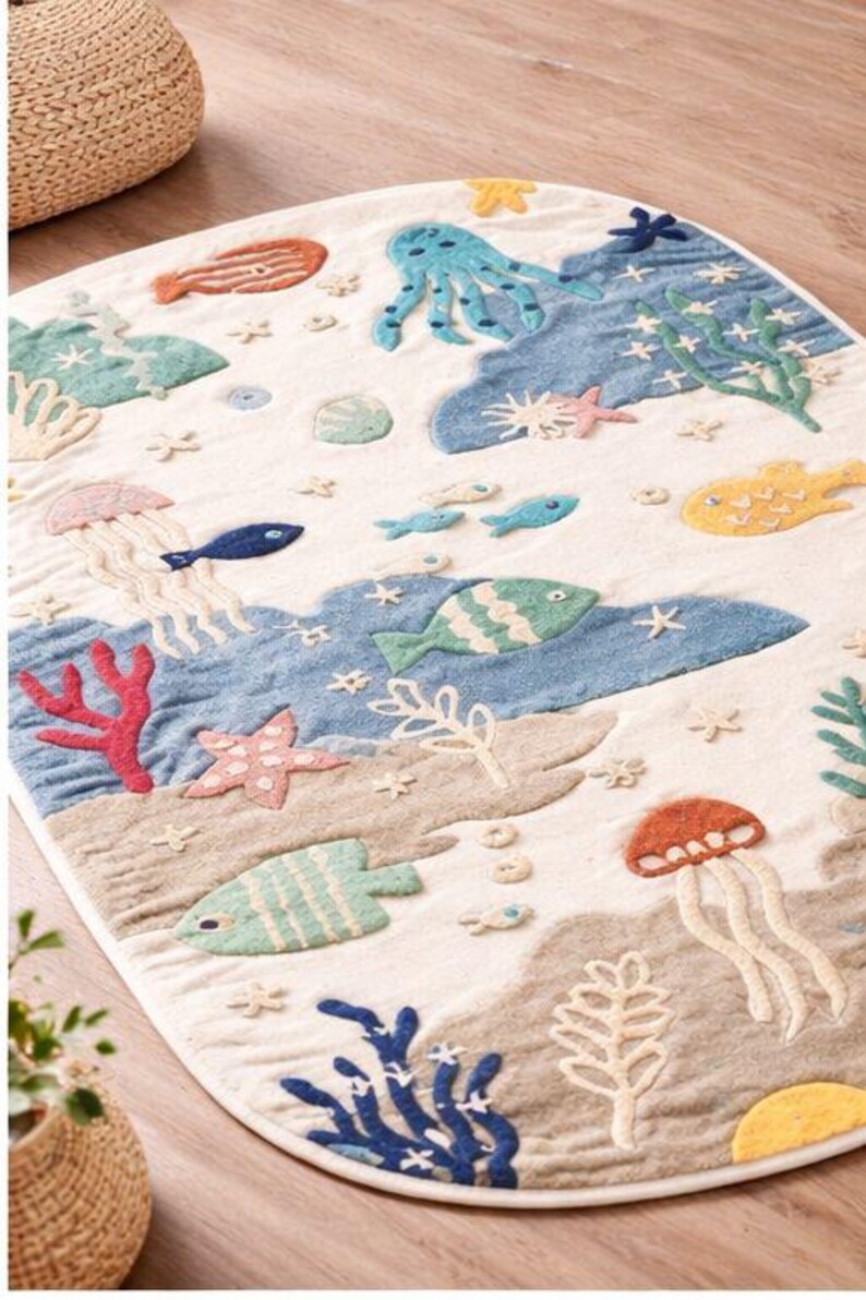 May include: An oval-shaped rug with a sea life design. The rug features various marine creatures, including fish, jellyfish, and an octopus, in shades of blue, green, orange, and yellow. The background is a sandy beige color, with blue water and white accents.