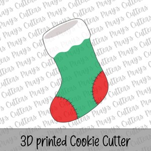 May include: A 3D printed cookie cutter shaped like a Christmas stocking. The design features a green body, a white cuff at the top, and a red toe and heel section. Black stitched detailing is visible on the red toe and heel areas.