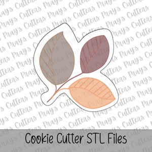 May include: A white cookie cutter with a three-leaf design featuring a grey leaf, a dark red leaf, and a light orange leaf. The leaves have thin, dark brown vein details. The text "Cookie Cutter STL Files" is printed at the bottom of the image.