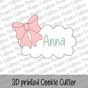 Puede incluir: Cortador de galletas impreso en 3D con forma de nube con un lazo rosa y el nombre "Anna" en verde. El texto "3D printed Cookie Cutter" está en la parte inferior de la imagen.