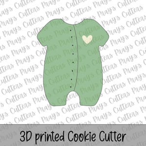 May include: A 3D-printed cookie cutter in the shape of a baby onesie. The onesie is light green with a white heart and black button details. The text "3D printed Cookie Cutter" is at the bottom.