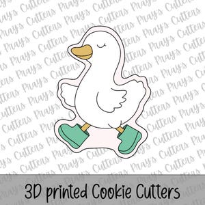 Può includere: Tagliapasta a forma di anatra bianca con becco giallo e stivali verde acqua. Il tagliapasta è bordato di nero e presenta il testo "3D printed Cookie Cutters" in basso.