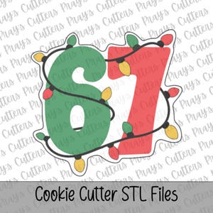 May include: A cookie cutter shaped like the number 67, featuring a green number 6 and a red number 7. The numbers are wrapped in a black string of Christmas lights with yellow and red bulbs. The design has a white outline and a white border.