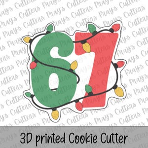 May include: A 3D printed cookie cutter shaped like the number 67, featuring a green number 6 and a red number 7. The design is wrapped in a black string of Christmas lights with small yellow and red bulbs, creating a festive holiday appearance.