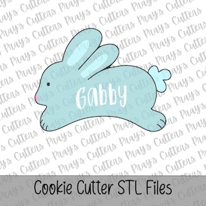 May include: A light blue bunny-shaped cookie cutter with the name "Gabby" in white script. The cutter has a small pink nose and a fluffy tail. The text "Cookie Cutter STL Files" is at the bottom.