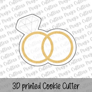 Puede incluir: Un cortador de galletas impreso en 3D con la forma de dos anillos de boda y un anillo de compromiso con diamantes. Los anillos son dorados y el diamante está delineado. El texto "3D printed Cookie Cutter" está en la parte inferior.