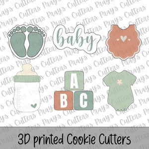 May include: A collection of 3D printed cookie cutters in the shape of baby-themed items. Includes a baby footprint, the word "baby", a bib, a baby bottle, ABC blocks, and a onesie. The cutters are in shades of green, orange, and white.