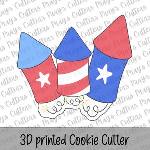 Pode incluir: Um cortador de biscoitos impresso em 3D com o formato de três foguetes de artifício. O foguete da esquerda é azul e vermelho com uma estrela branca, o central tem listras azuis e brancas, e o da direita é vermelho e azul com uma estrela branca.