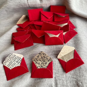 May include: A collection of small, red paper envelopes, some open to reveal patterned or printed inserts. The envelopes are scattered on a neutral-toned fabric surface, creating a visually appealing arrangement.