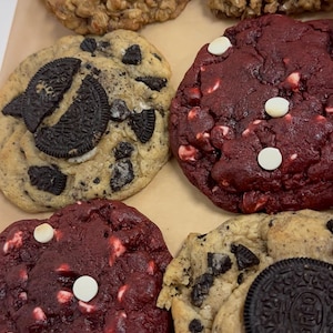 May include: An assortment of freshly baked cookies in a box. The cookies include oatmeal with almond slices, red velvet with white chocolate chips, and cookies and cream with Oreo cookie pieces.