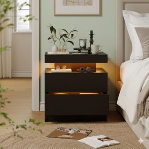 May include: A modern black nightstand with two drawers and a shelf with warm lighting. The top surface displays a vase with flowers, a candle, and a mug. A pair of glasses and headphones rest on the shelf. The nightstand is next to a bed.