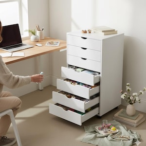 Peut inclure: Un meuble de rangement blanc à cinq tiroirs, avec des tiroirs ouverts révélant des fournitures d'artisanat. Le meuble est sur roulettes et se trouve à côté d'un bureau en bois avec un ordinateur portable. Une personne est assise près du bureau.